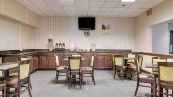 Habitación de hotel con kitchenette, televisor de pantalla plana montado en la pared y comedor con mesas y sillas.