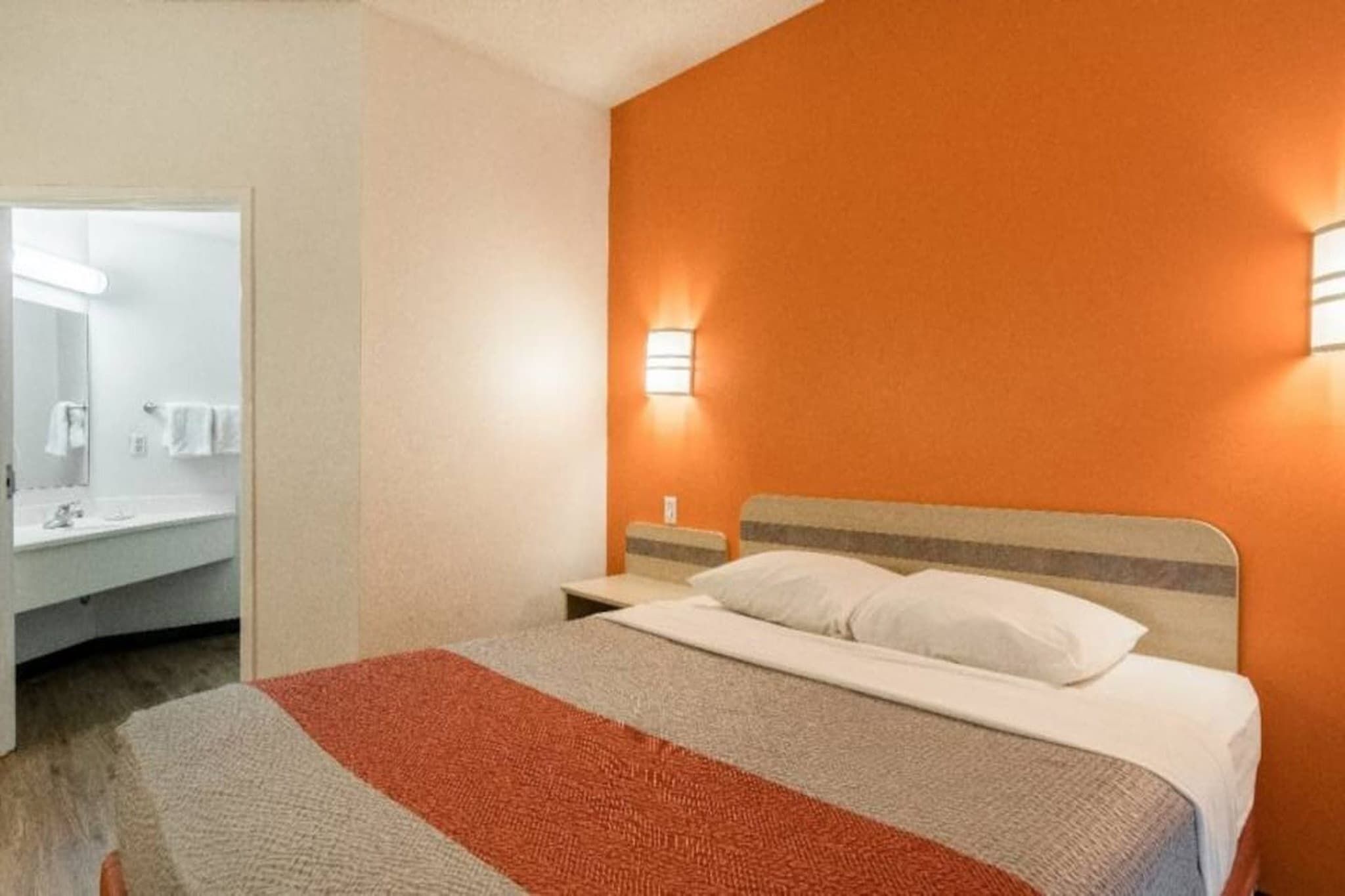 A hotel room with a bed, a nightstand, a wall-mounted lamp, and a bathroom with a mirror and towel rack.