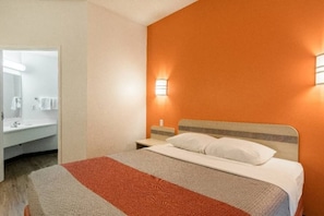Habitació d'hotel amb llit, una petita taula de nit, una làmpada penjada a la paret i un bany amb mirall i barra de tovalloles.