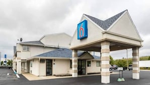 Un motel con un letrero que dice '6', un área de estacionamiento techada y un edificio de una sola planta.