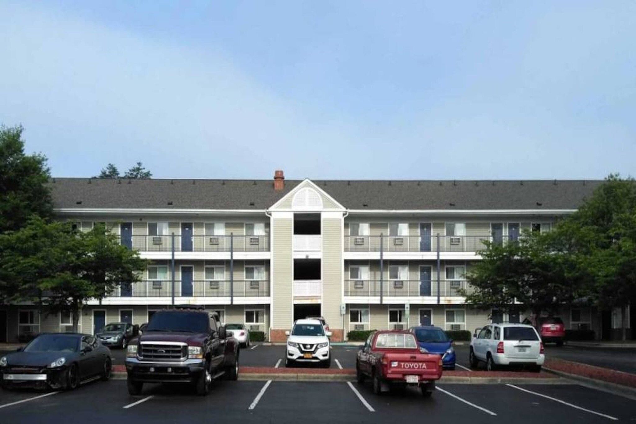 A multi-story building with a central entrance, parking lot with several cars, and a symmetrical facade with windows and a flat roof.