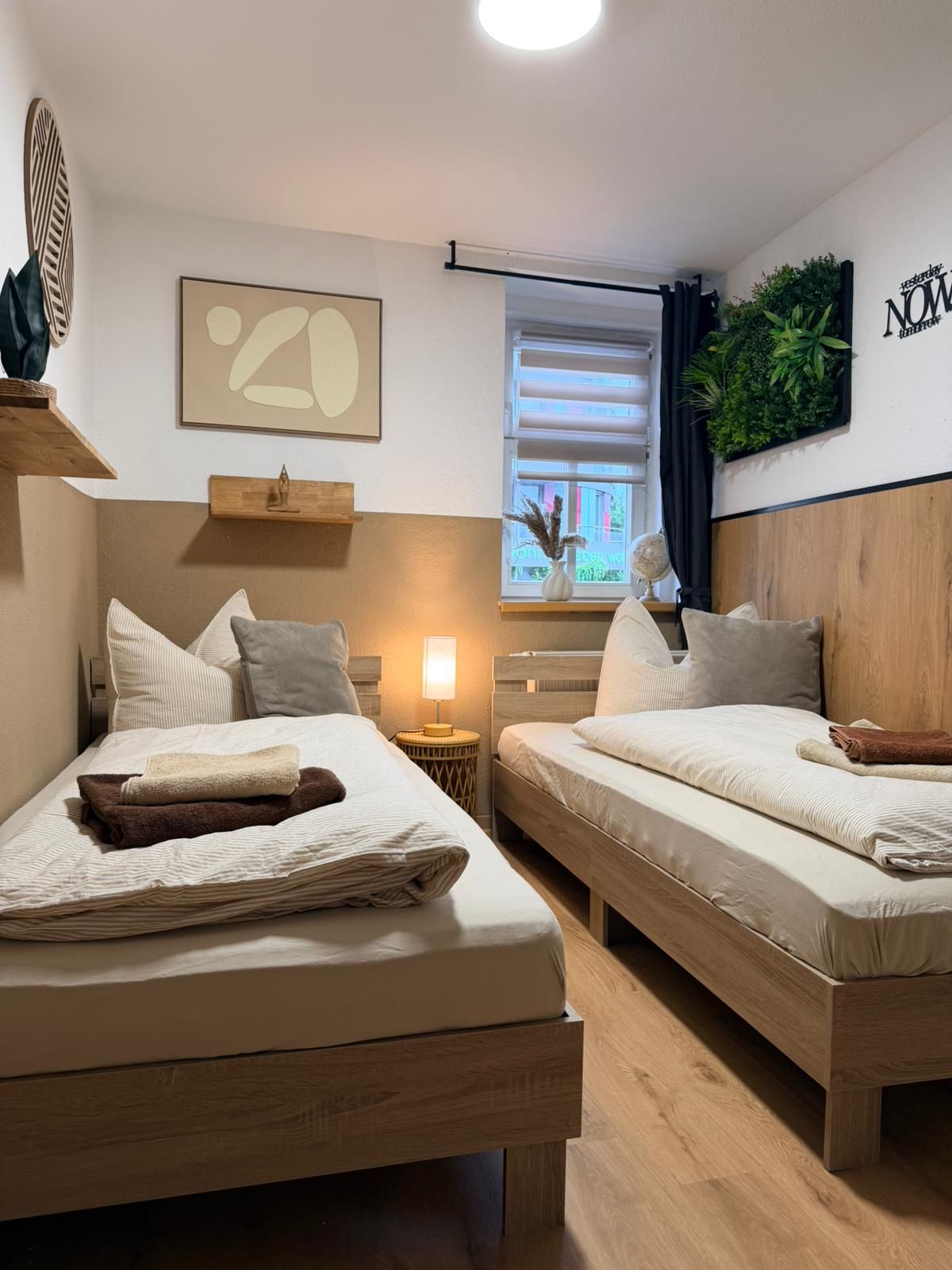 Ein Hotelzimmer mit zwei Betten, einem Fenster mit Jalousien, einer Wandpflanze und einem gerahmten abstrakten Kunstwerk.