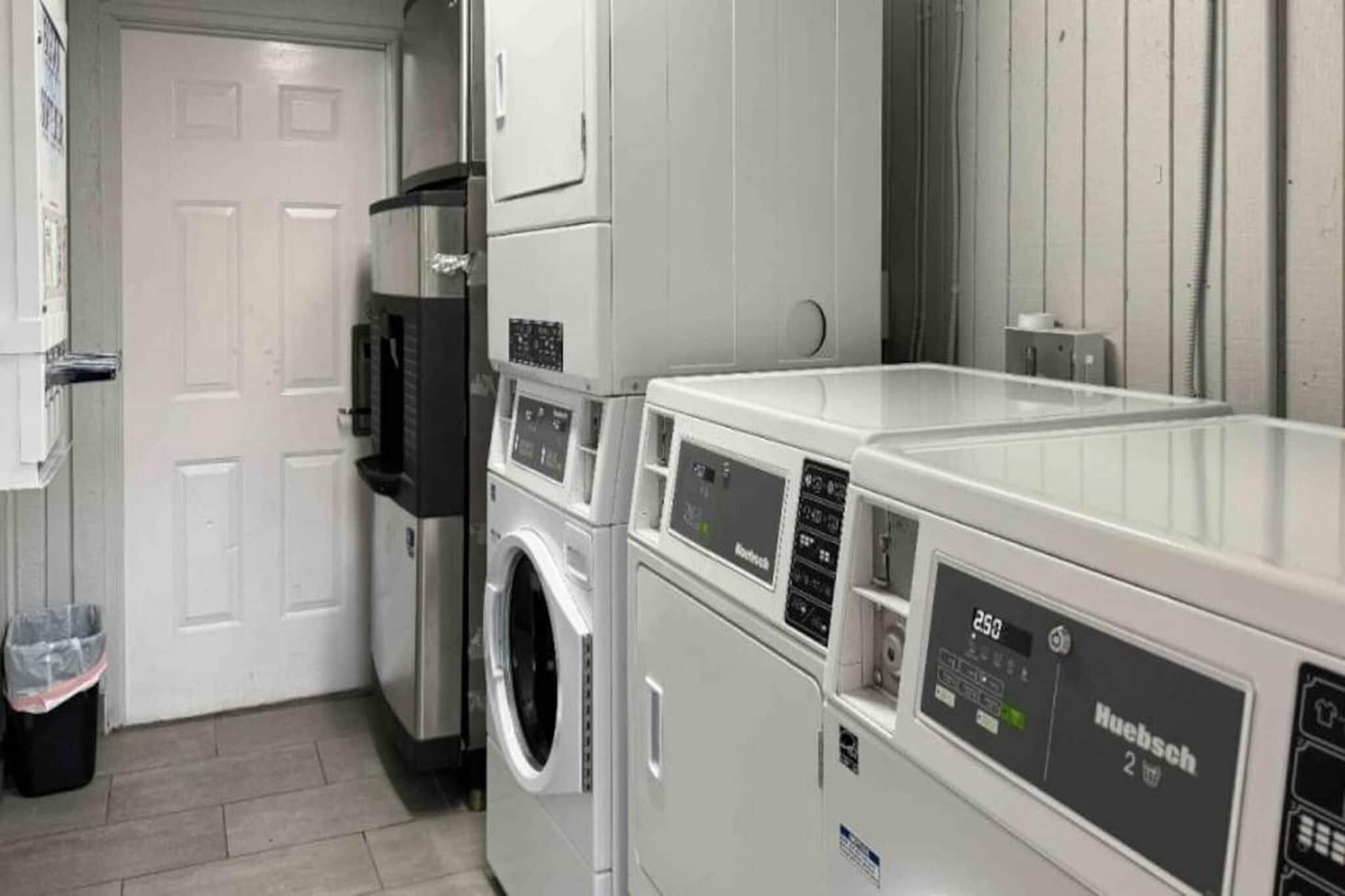 A laundry room with a front-loading washer and dryer, a trash bin, and a door leading to another room.