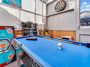 Une table de billard, un jeu de fléchettes et un billard d’arcade se trouvent dans une pièce aux murs en bois et dotée d’une fenêtre.