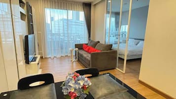 A modern hotel room with a sofa, a dining table with chairs, a TV, and a large window with a city view.