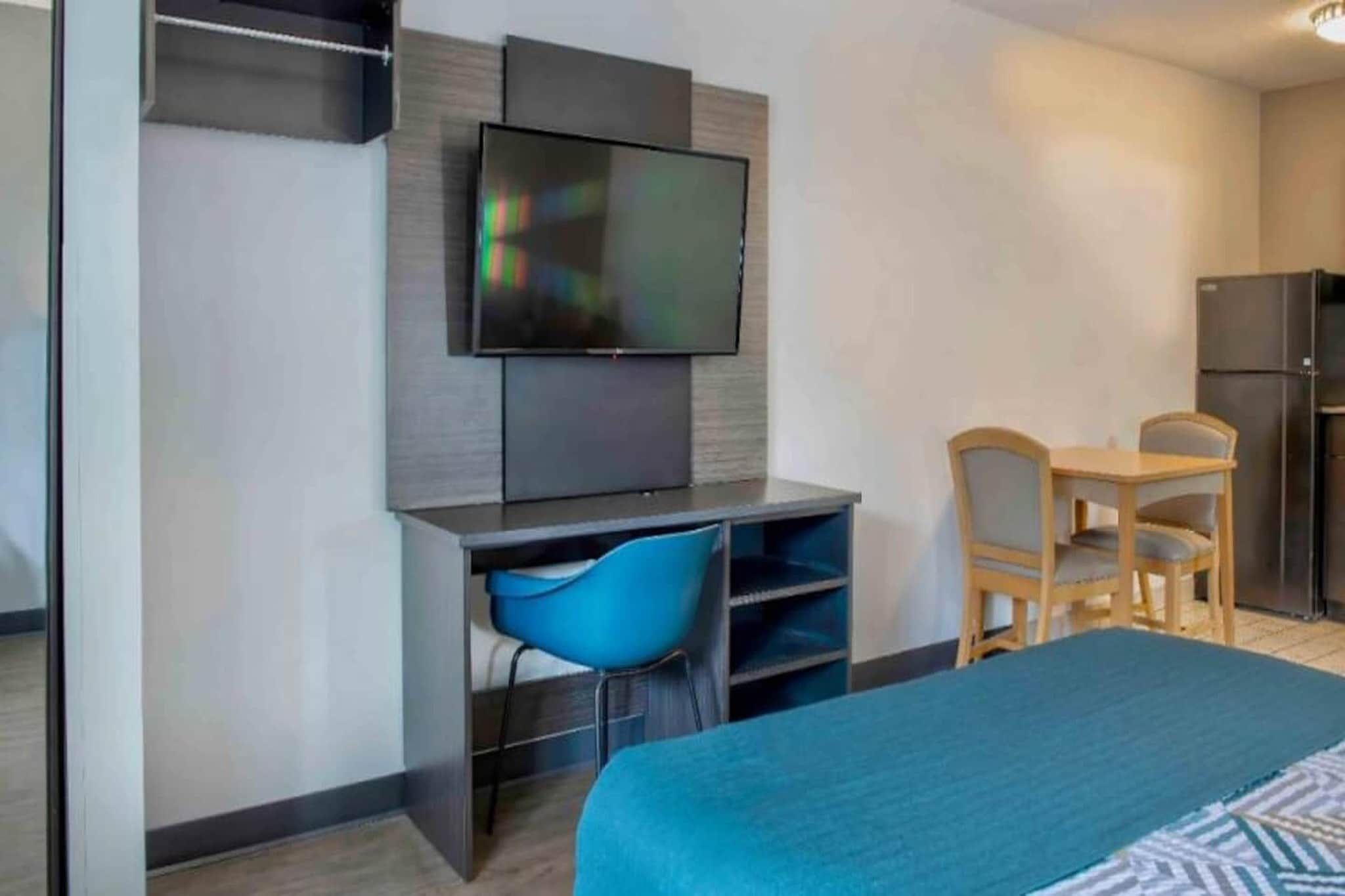 Una habitación de hotel con cama, escritorio con silla, televisor montado en la pared y refrigerador.