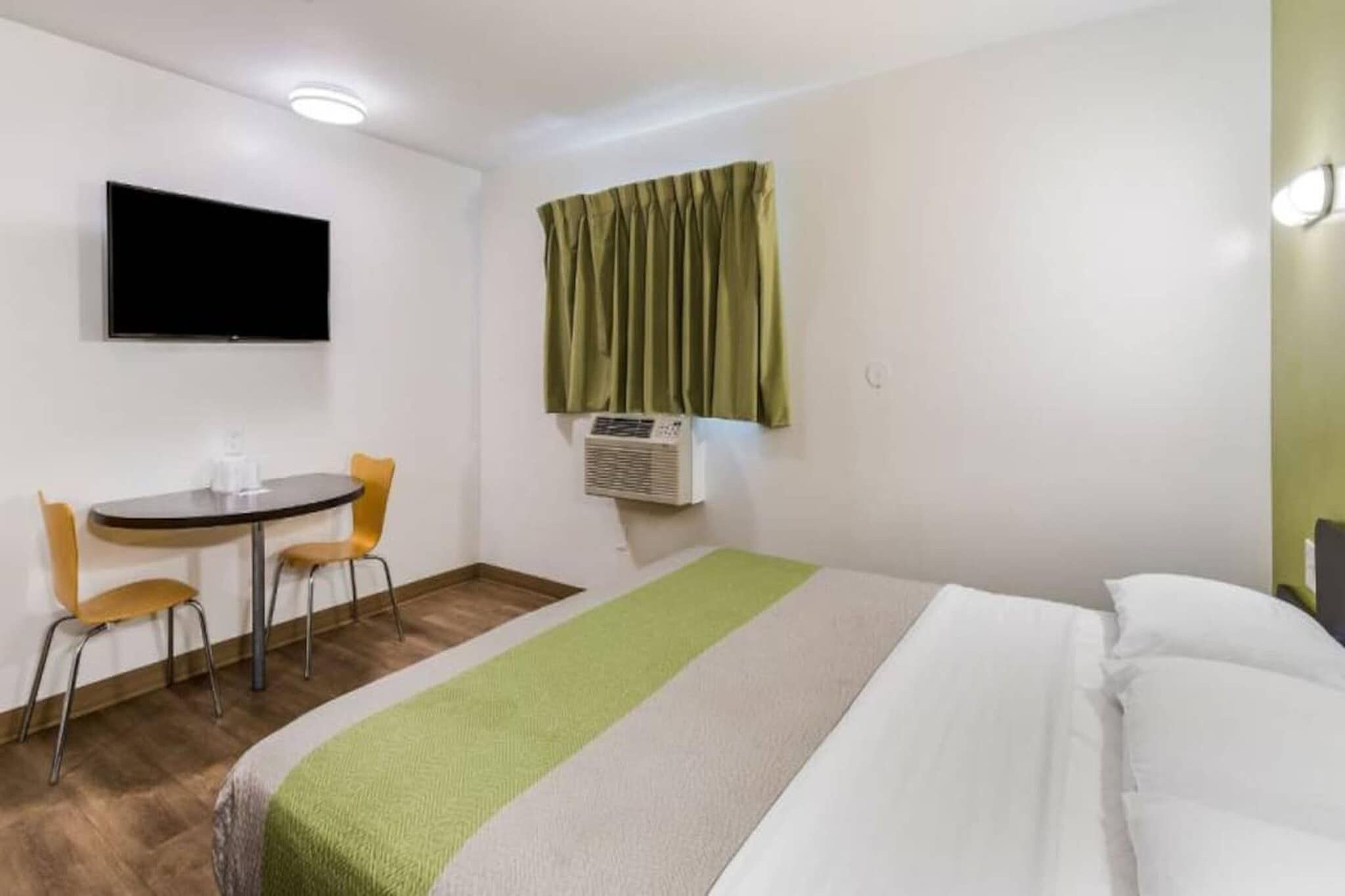 A hotel room with a bed, a desk, two chairs, a television, and an air conditioning unit.