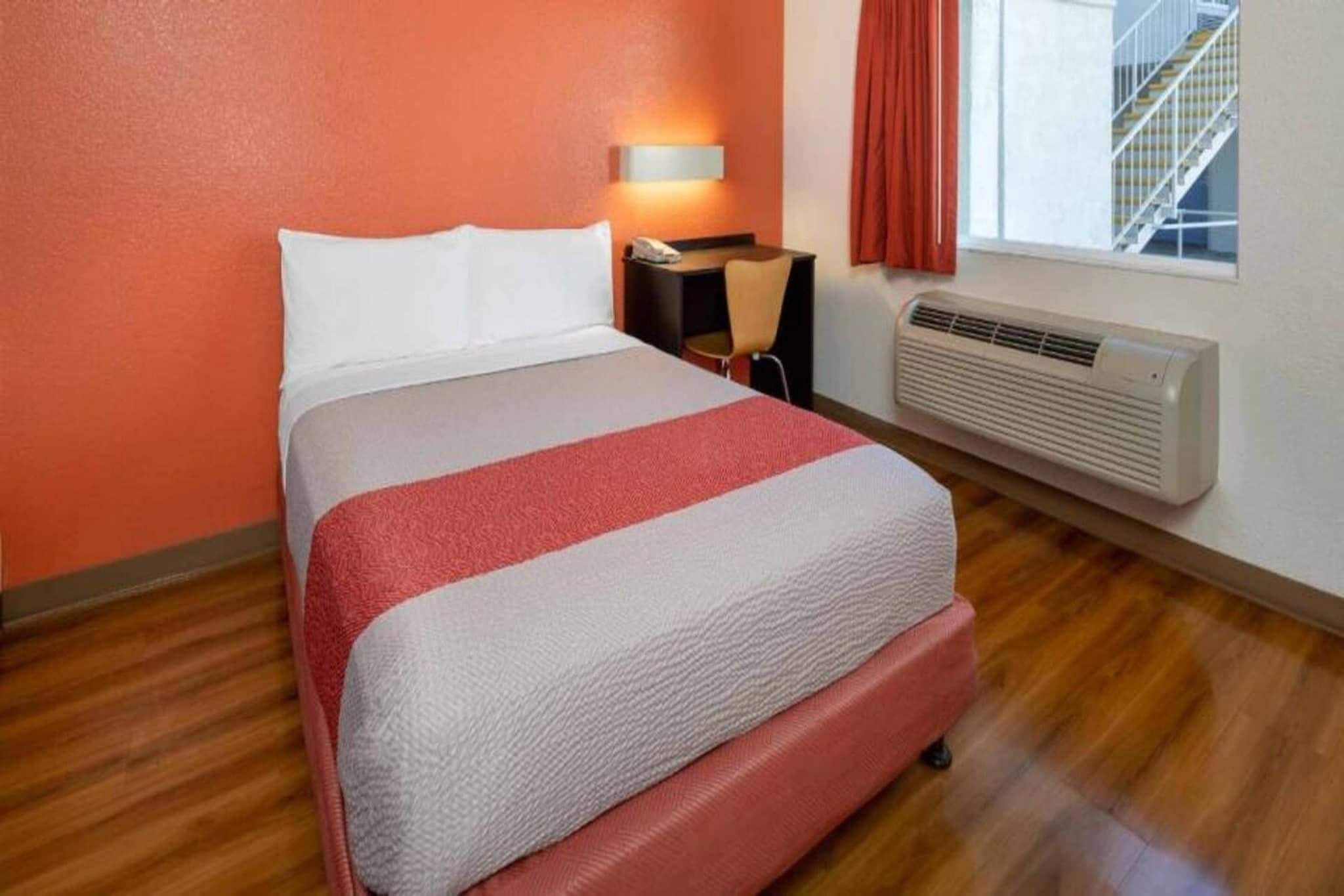 Ein Hotelzimmer mit einem Bett, einem Nachttisch mit Lampe, einem Fenster mit Blick auf eine Treppe und einer Wandklimaanlage.