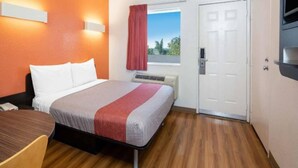 Quarto de hotel com uma cama, escrivaninha, cadeira, janela com cortinas e porta.