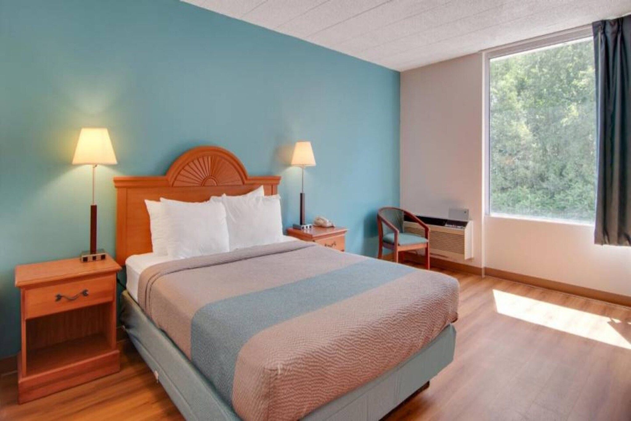 Une chambre d’hôtel avec un lit, deux tables de chevet, une chaise, un bureau et une fenêtre avec des rideaux.