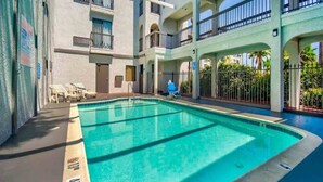 Una piscina con superficie de agua azul cristalina, rodeada por un edificio con varios balcones y un paseo cubierto.
