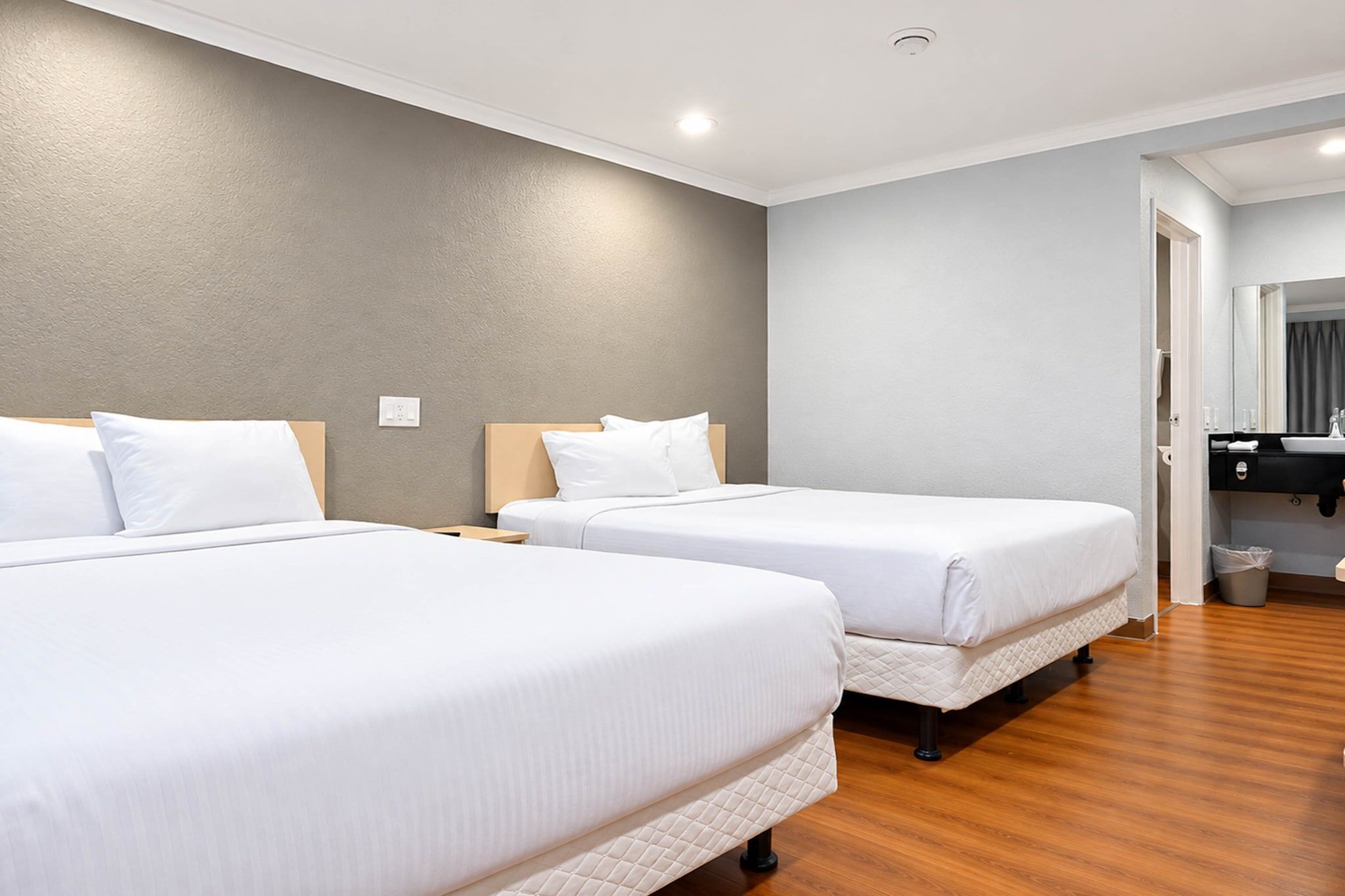 A hotel room with two beds, a wooden floor, a grey wall, and a bathroom visible through an open door.