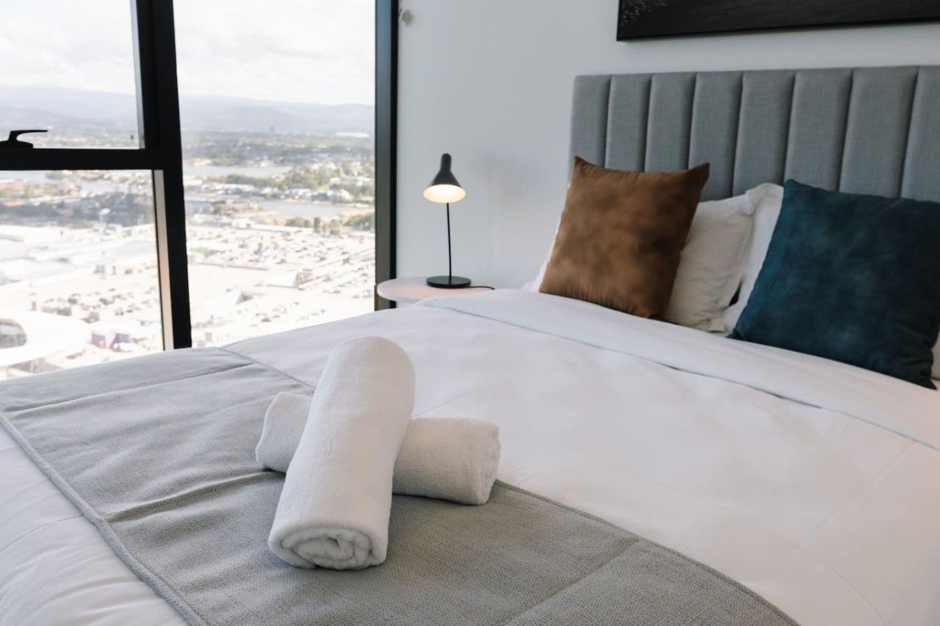 Una cama bien hecha con una toalla enrollada, un cabecero gris y almohadas de varios colores.
