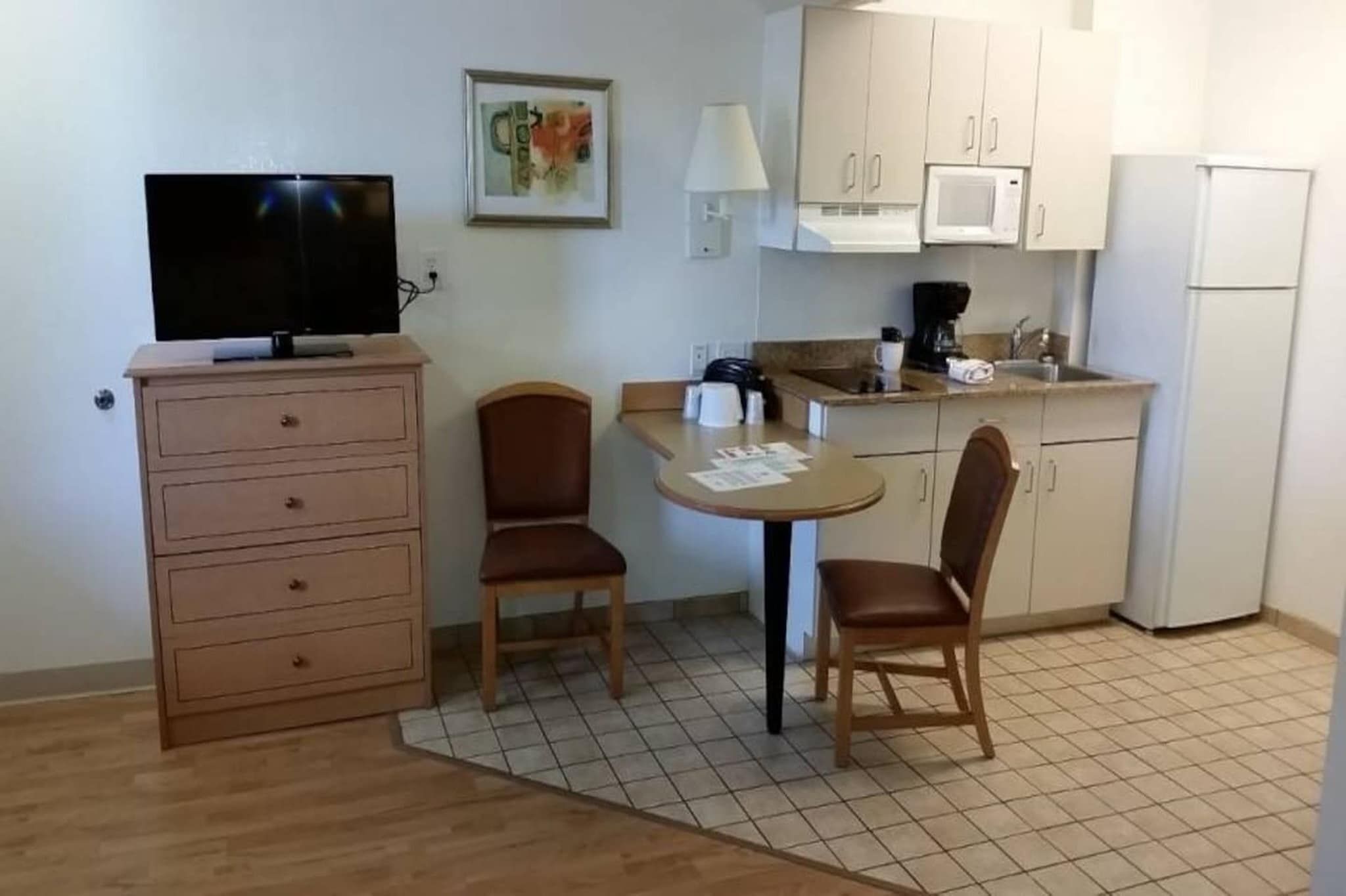 Ett litet pentry med mikrovågsugn, kaffebryggare och kylskåp. Där finns ett träbord med två stolar och en tv-apparat placerad på en byrå.