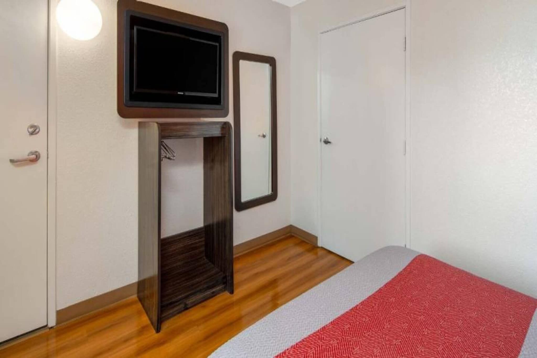 Une pièce avec un plancher en bois, un tapis rouge et blanc, un téléviseur fixé sur un meuble en bois, une porte et un miroir au mur.