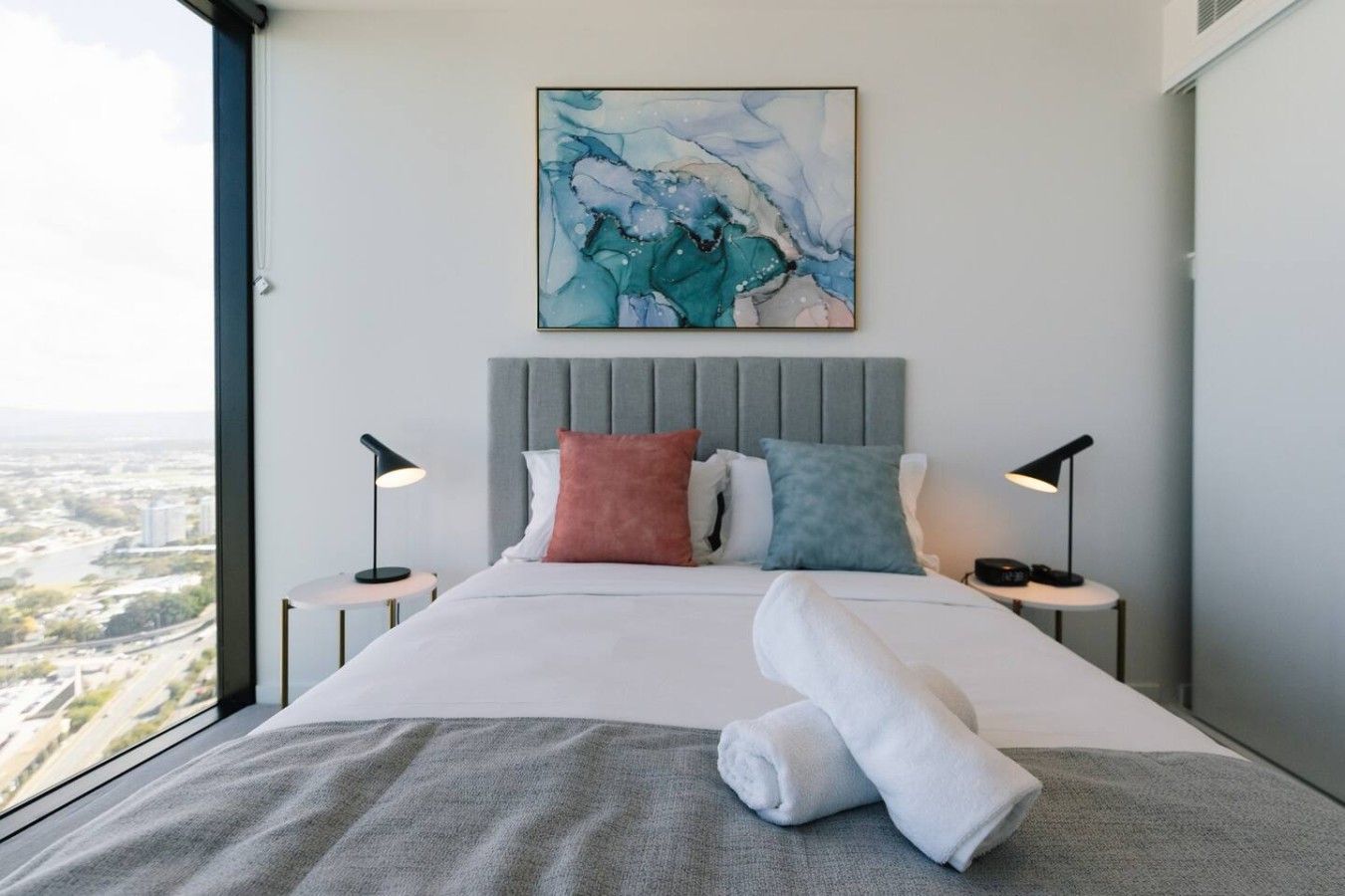 A modern hotel room with a large bed, two bedside tables, a gray headboard, and a city view.