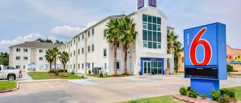 Un motel con un llamativo letrero con el número '6', un estacionamiento y un edificio con varias ventanas y techo plano.