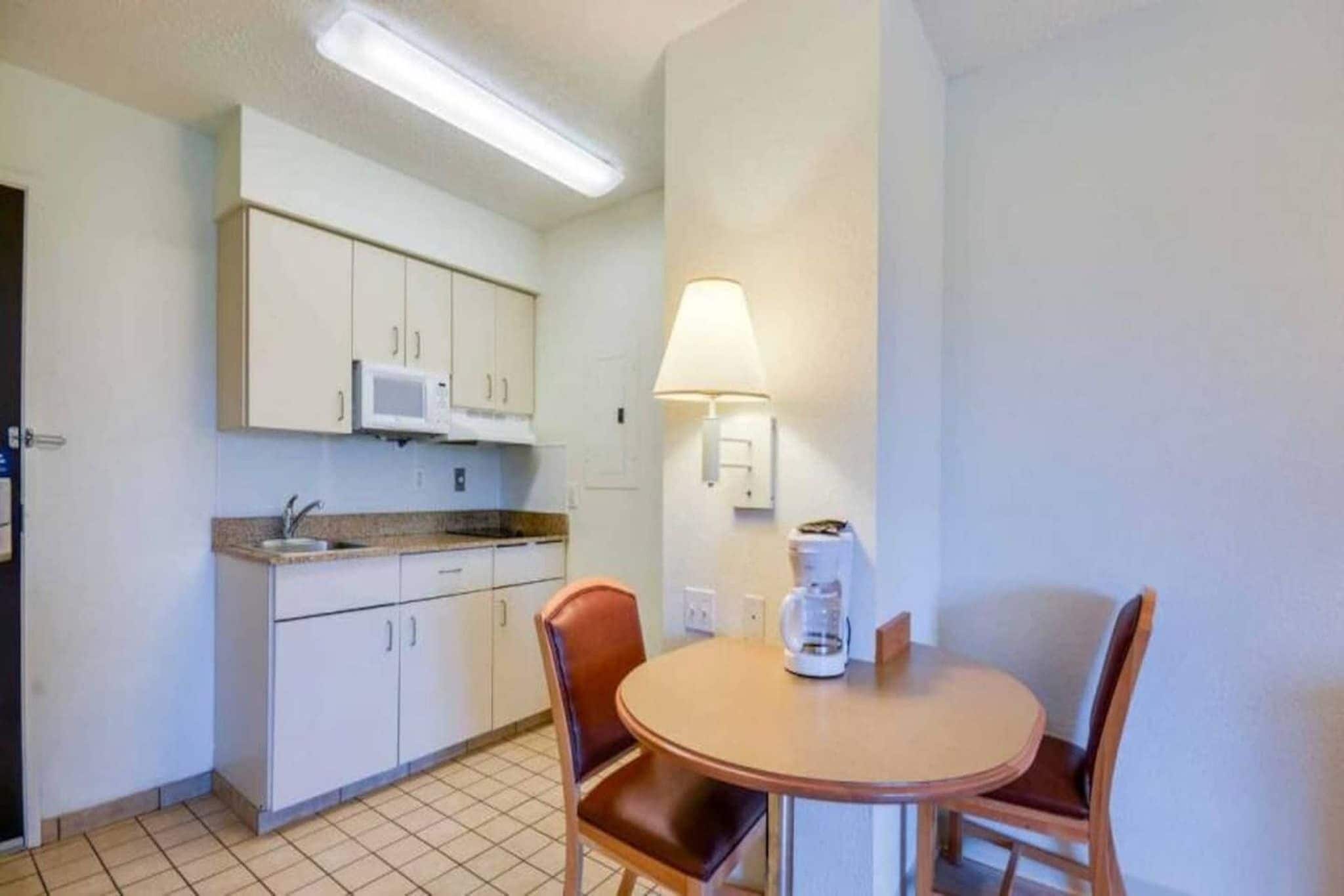 Une cuisine compacte avec des meubles blancs, un four à micro-ondes, une machine à café posée sur la table et une lampe fixée au mur.
