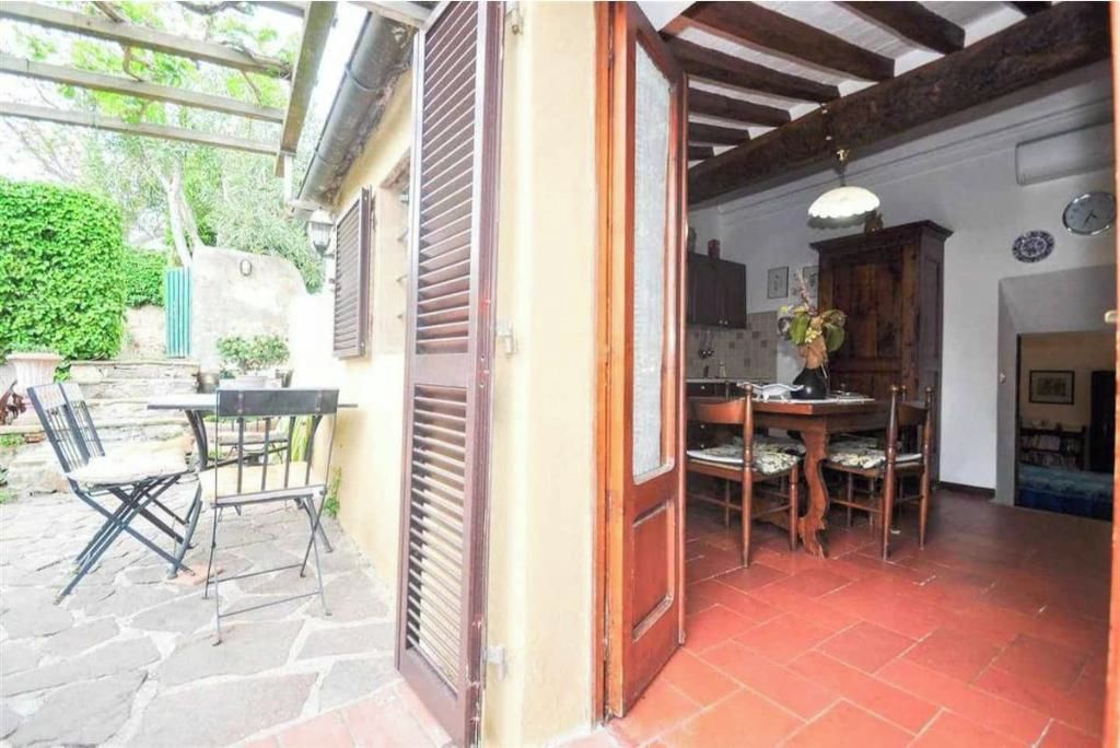 Une terrasse ouverte avec un sol en pierre, une table en bois entourée de chaises et une porte donnant sur un espace intérieur doté d’un sol carrelé et d’un luminaire suspendu.
