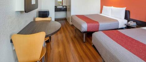 Habitación de hotel con piso de madera, una mesa de comedor redonda, dos camas individuales con ropa de cama roja y gris, una kitchenette pequeña y un televisor montado en la pared.
