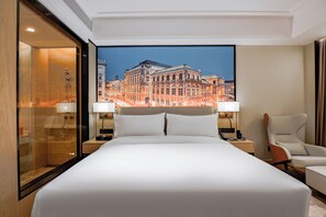 Une chambre d’hôtel avec un grand lit, deux tables de chevet, une chaise et une grande fenêtre donnant sur un bâtiment historique.