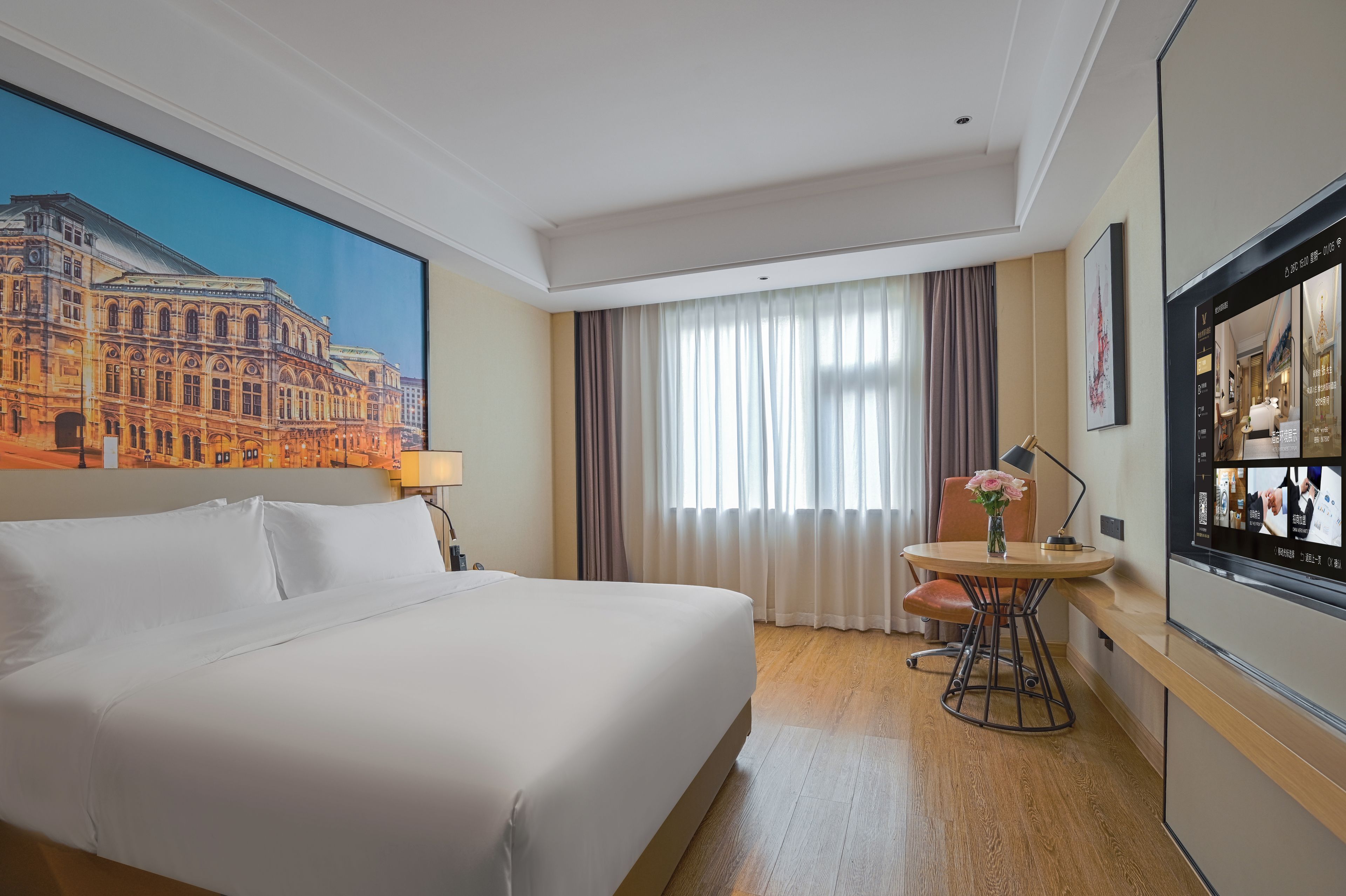 A hotel room with a large bed, a desk with a chair, a window with curtains, and a painting of a building on the wall.