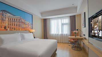 A hotel room with a large bed, a desk with a chair, a window with curtains, and a painting of a building on the wall.