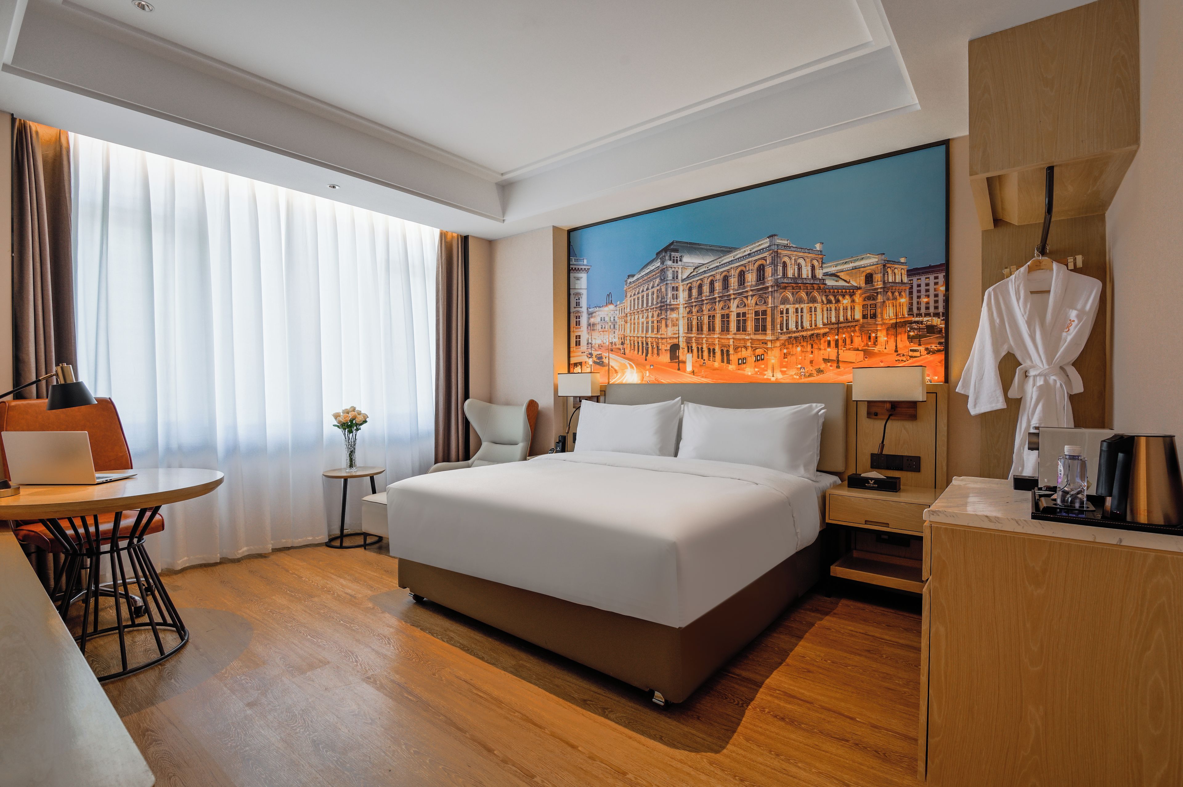 Habitación de hotel con una cama grande, un escritorio con una computadora portátil, una silla, una mesita con un florero, un cuadro de un paisaje urbano y una bata colgada en la pared.