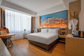 A hotel room with a large bed, a desk with a laptop, a chair, a small table with a vase of flowers, a painting of a cityscape, and a robe hanging on the wall. - Vienna International Hotel  (Shenzhen)