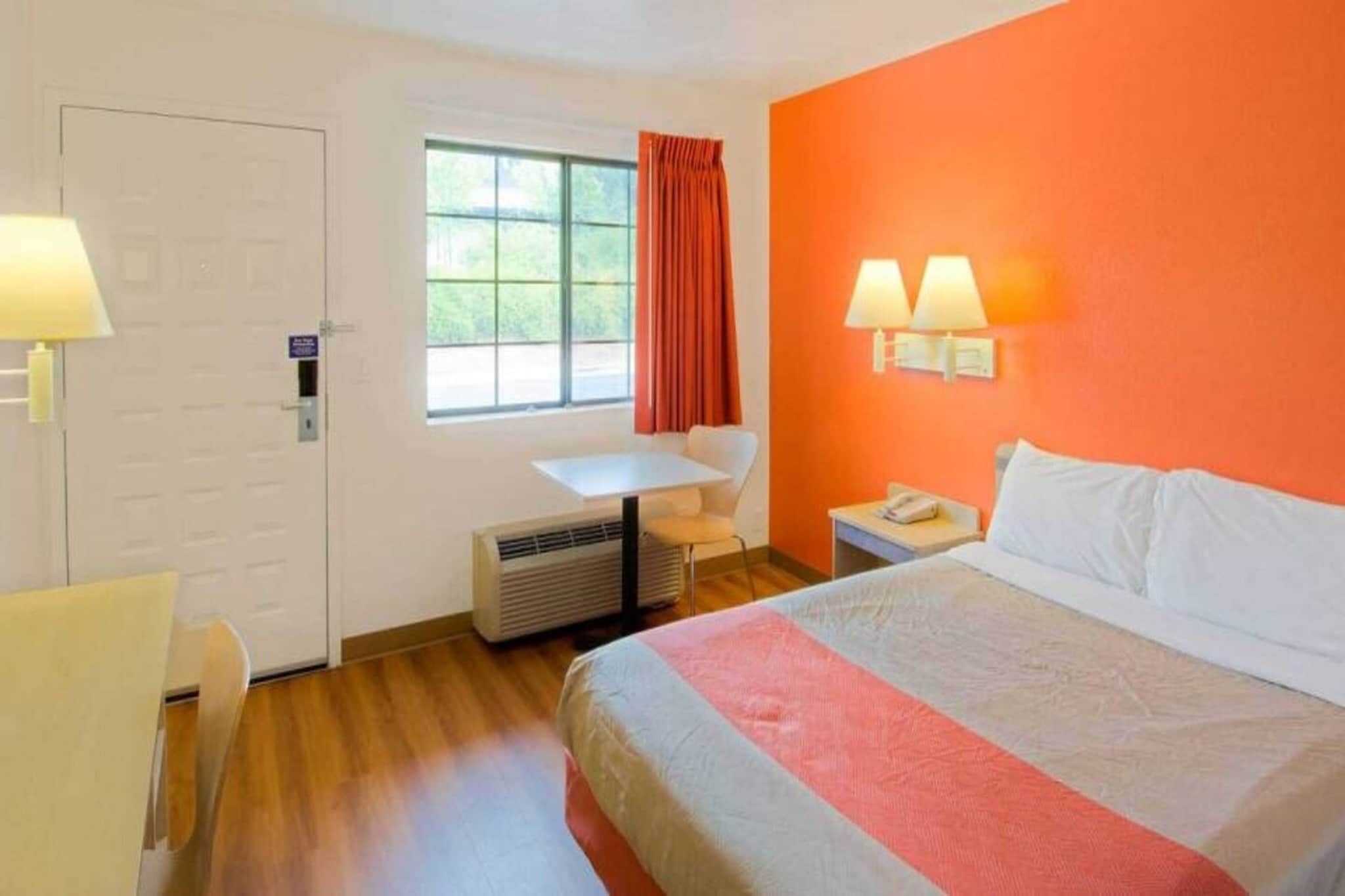 A hotel room with a bed, a desk, a chair, a window with curtains, and a wall-mounted lamp.