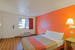Habitación de hotel con cama, ventana con cortinas, unidad de aire acondicionado, escritorio con lámpara y puerta.