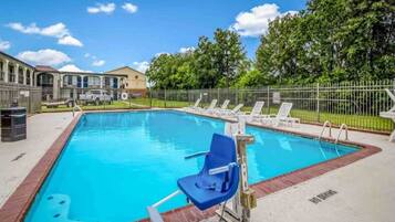 A swimming pool with lounge chairs, a lifeguard stand, and a fenced area with a building in the background.
