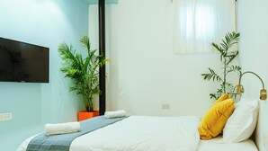 Habitación de hotel con una cama, televisor montado en la pared, una planta en maceta y una mesita de noche con lámpara.