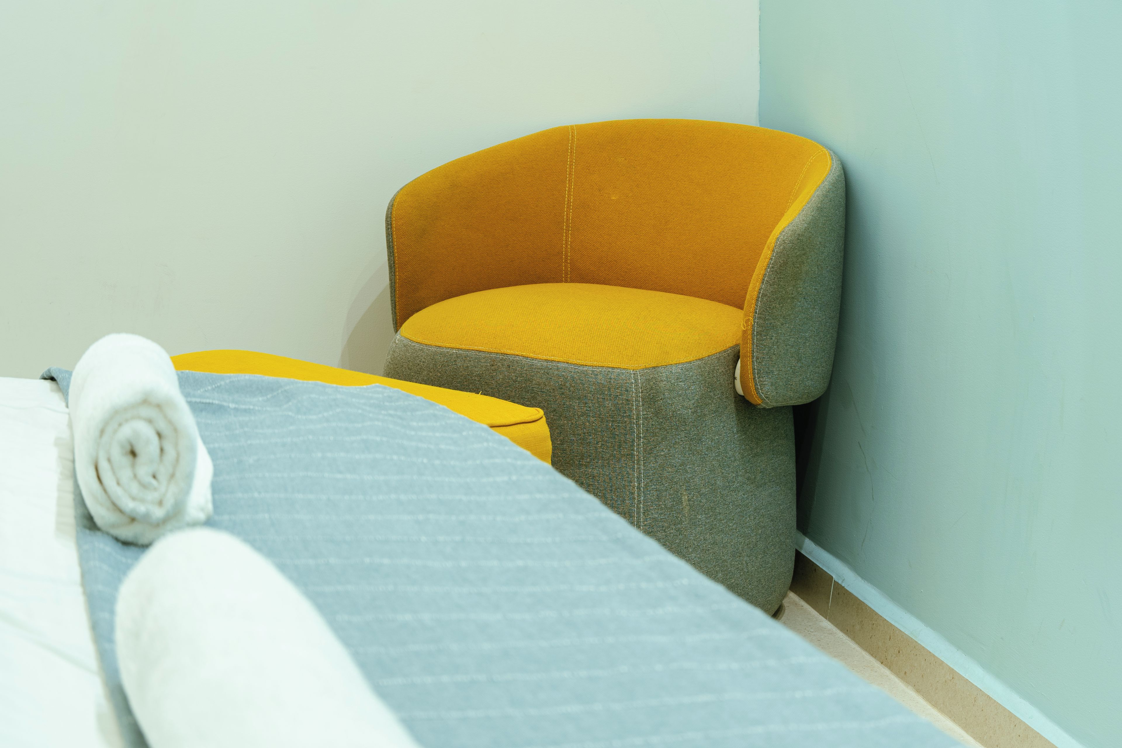 A room with two beds, one with a rolled white towel and the other with a grey blanket.