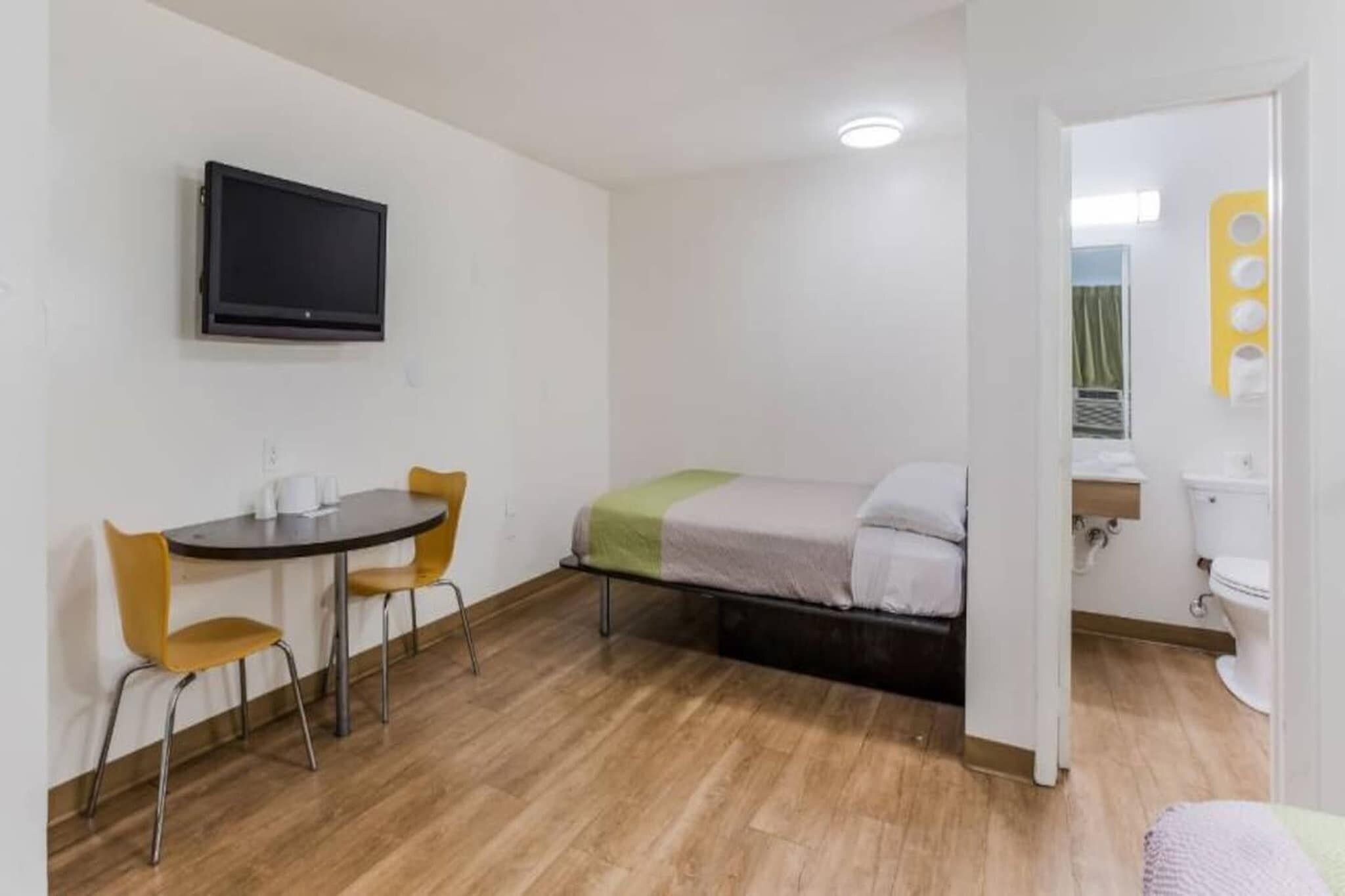Een kleine kamer met een bed, een bureau met twee stoelen, een aan de muur bevestigde televisie, en een badkamer zichtbaar door een geopende deur.