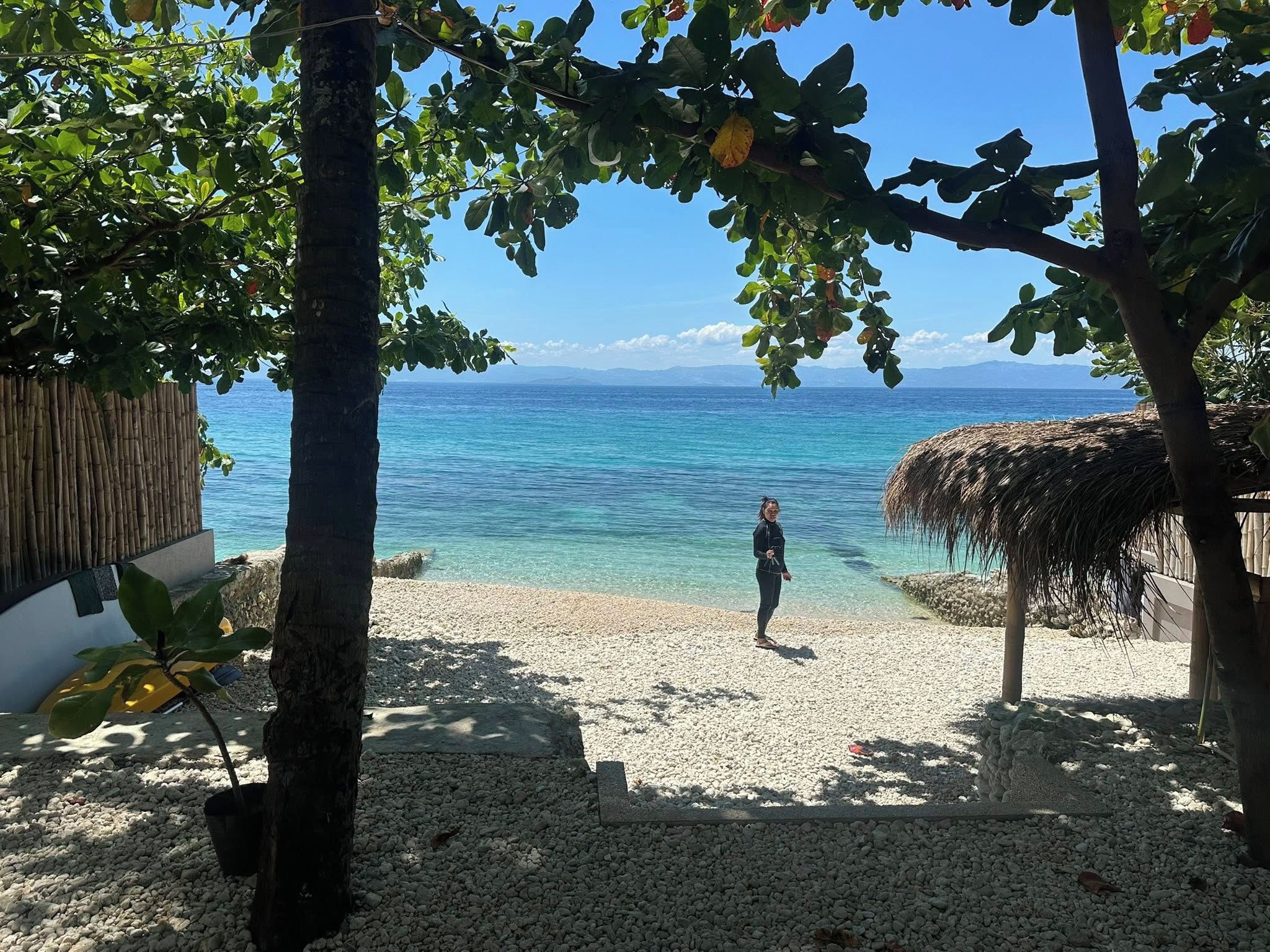 A beach with clear water, a person standing, and a thatched-roof structure.