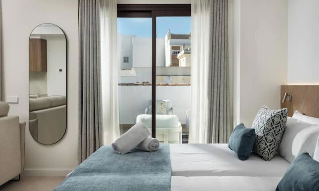 Una habitación de hotel con una cama, un espejo, una ventana con cortinas y vistas a edificios.