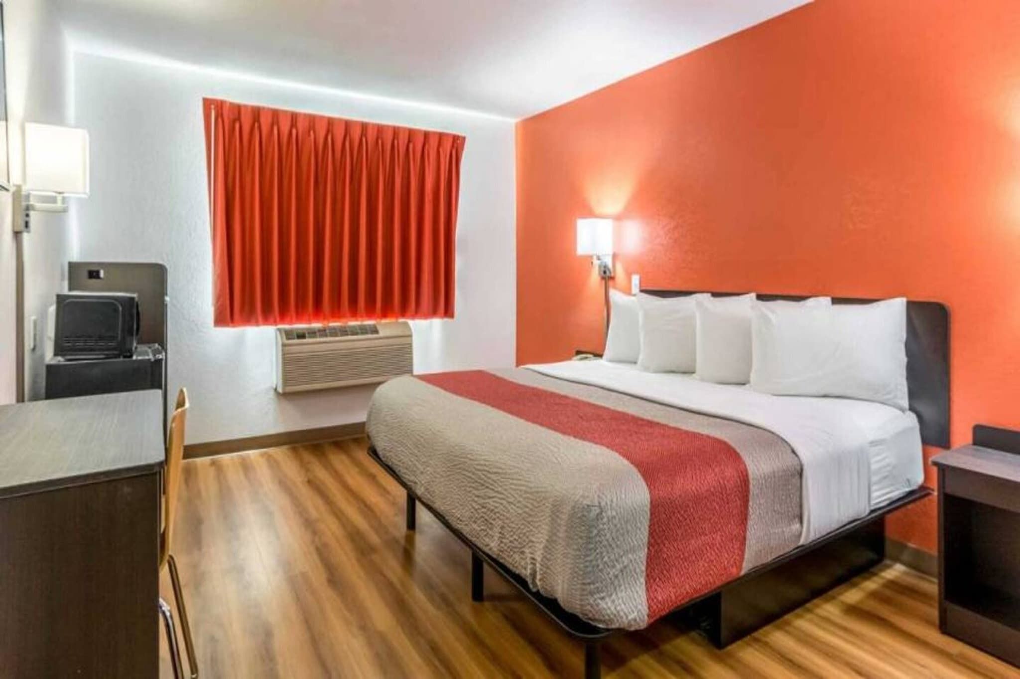 A hotel room with a bed, a desk, a chair, a window with curtains, an air conditioner, and a wall-mounted lamp.