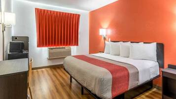 A hotel room with a bed, a desk, a chair, a window with curtains, an air conditioner, and a wall-mounted lamp.