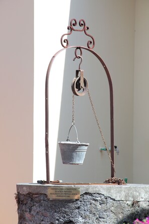 A historical water collection cistern with a metal bucket suspended by a chain, mounted on a stone base with a plaque.