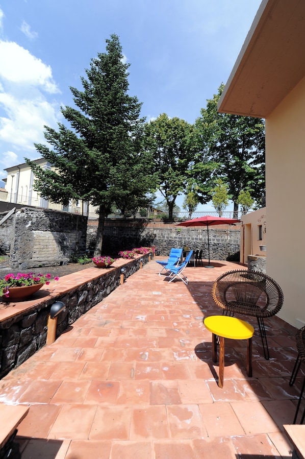 A patio area with terracotta tiles, a stone wall, a large tree, and outdoor furniture including a yellow table and blue chairs.