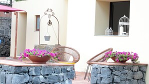 A patio with stone walls, two wicker chairs, and flower pots with pink flowers. - Vrbo Property (Fornazzo)