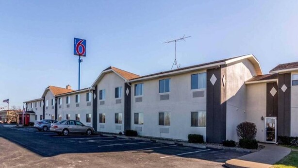 Un motel reconnaissable à son grand panneau avec le chiffre « 6 », un parking et une rangée de bâtiments à un seul étage, caractérisés par une architecture simple.