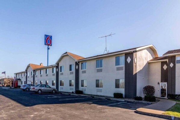 A motel with a prominent '6' sign, a parking lot, and a row of single-story buildings with a simple architectural design. - 2 Queen Beds | Smoking (HARRISVILLE)