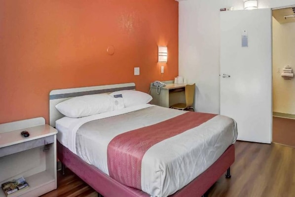 A single-bed room with a wooden floor, a white and red bedspread, a bedside table with a lamp, and a small desk with a telephone.