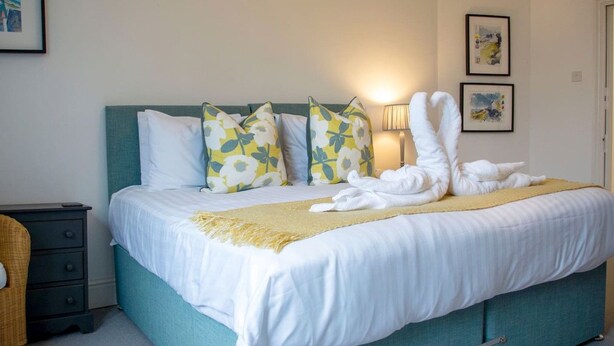 A neatly made bed with white bedding and floral-patterned pillows, a bedside lamp, and a small bedside table with a lamp. - Malvern Hills 3-Bed Retreat | Sleeps 8 | Free Park (Worcestershire)