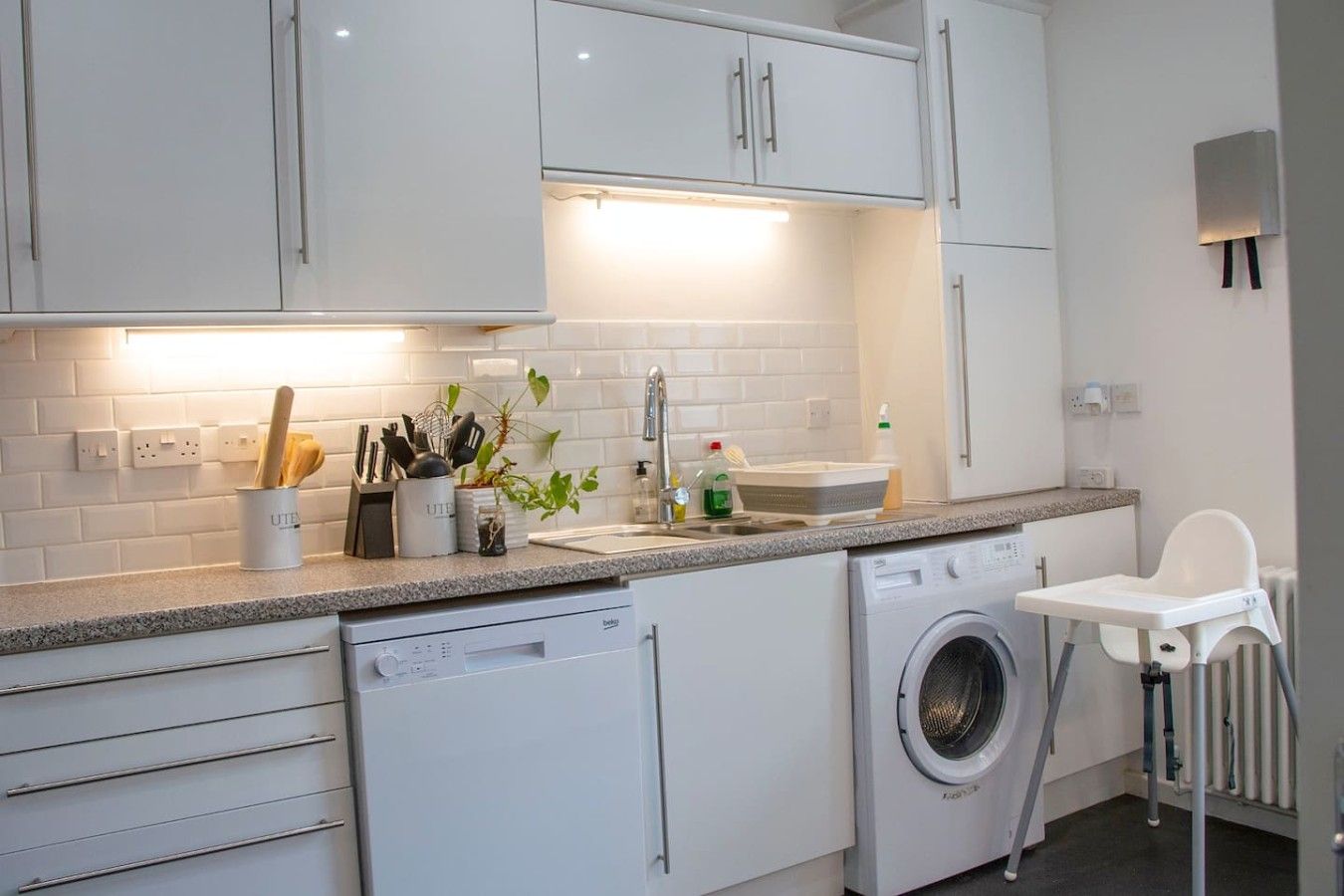 Een moderne keuken met witte kasten, een vaatwasser, een wasmachine en een kinderstoel.