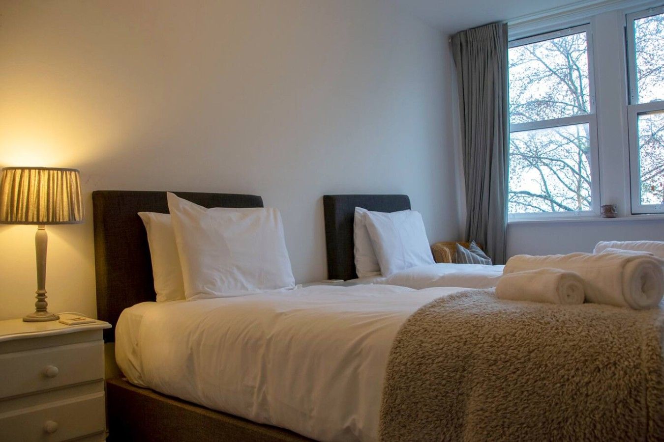 Une chambre d’hôtel avec deux lits, une lampe de chevet, une fenêtre avec des rideaux et une vue sur les arbres.