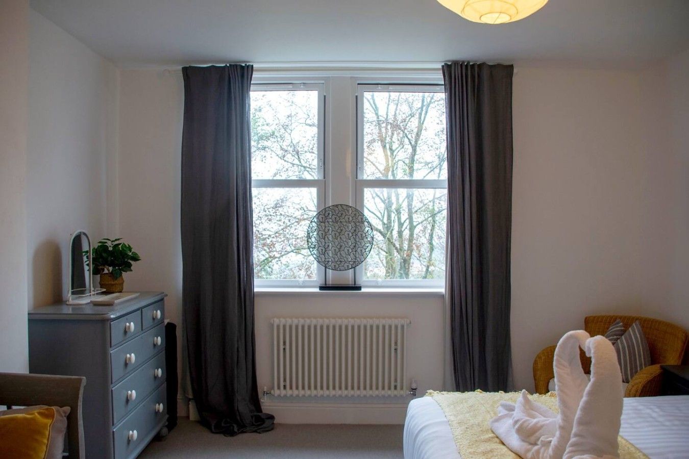 Une chambre à coucher avec un lit, une commode, un radiateur et une fenêtre avec des rideaux.