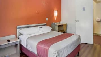 A single-bed room with a wooden floor, a white and red bedspread, a bedside table with a lamp, and a small desk with a telephone.